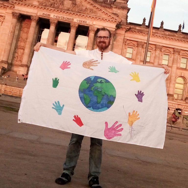 Mann hält eine Flagge mit bunten Handabdrücken und der Erde vor dem Reichstag.